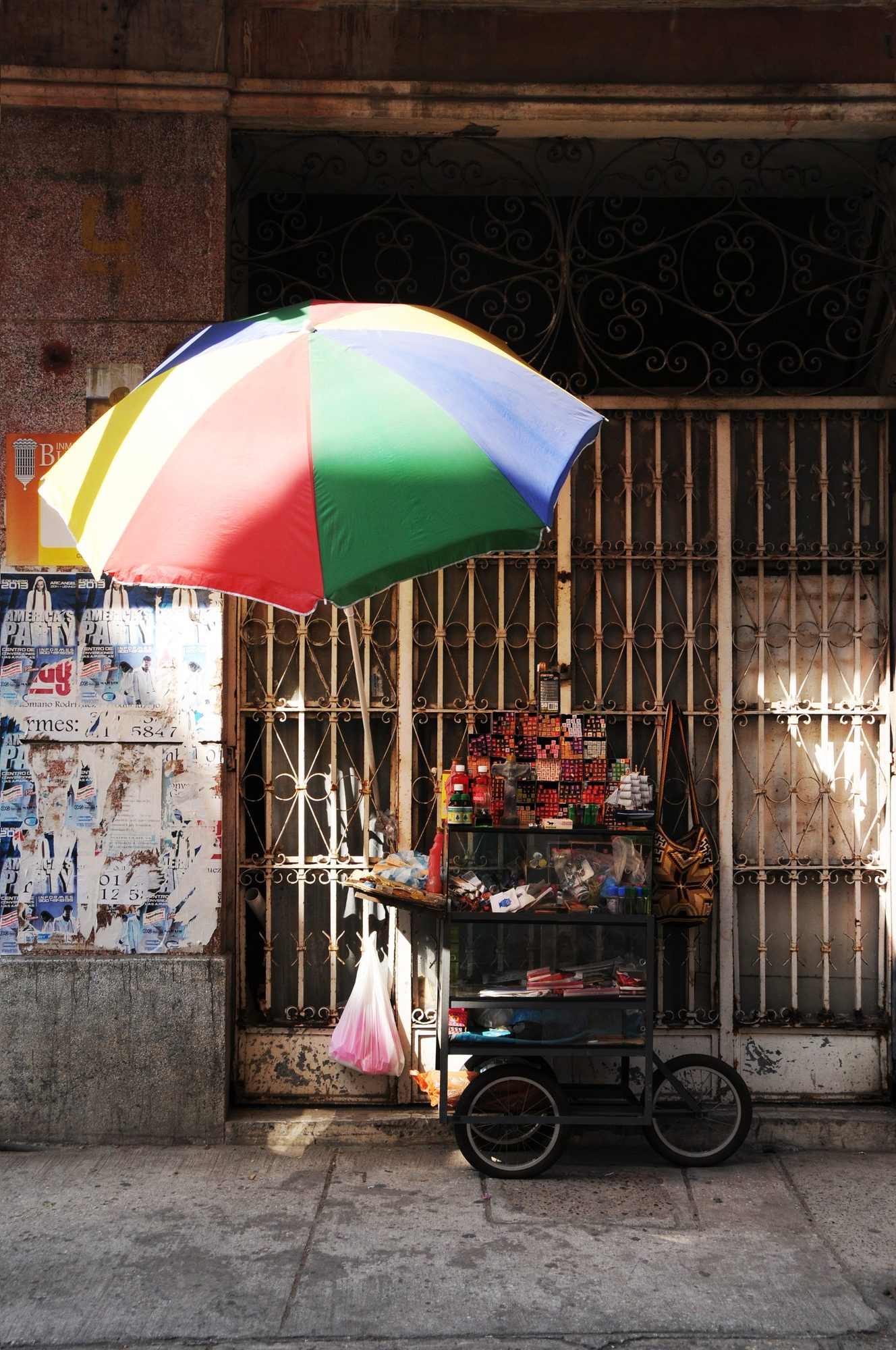 A vendor's cart shaded by a rainbow umbrella outside a shuttered doorway, Cartagena, Colombia — photograph by Brian Walker