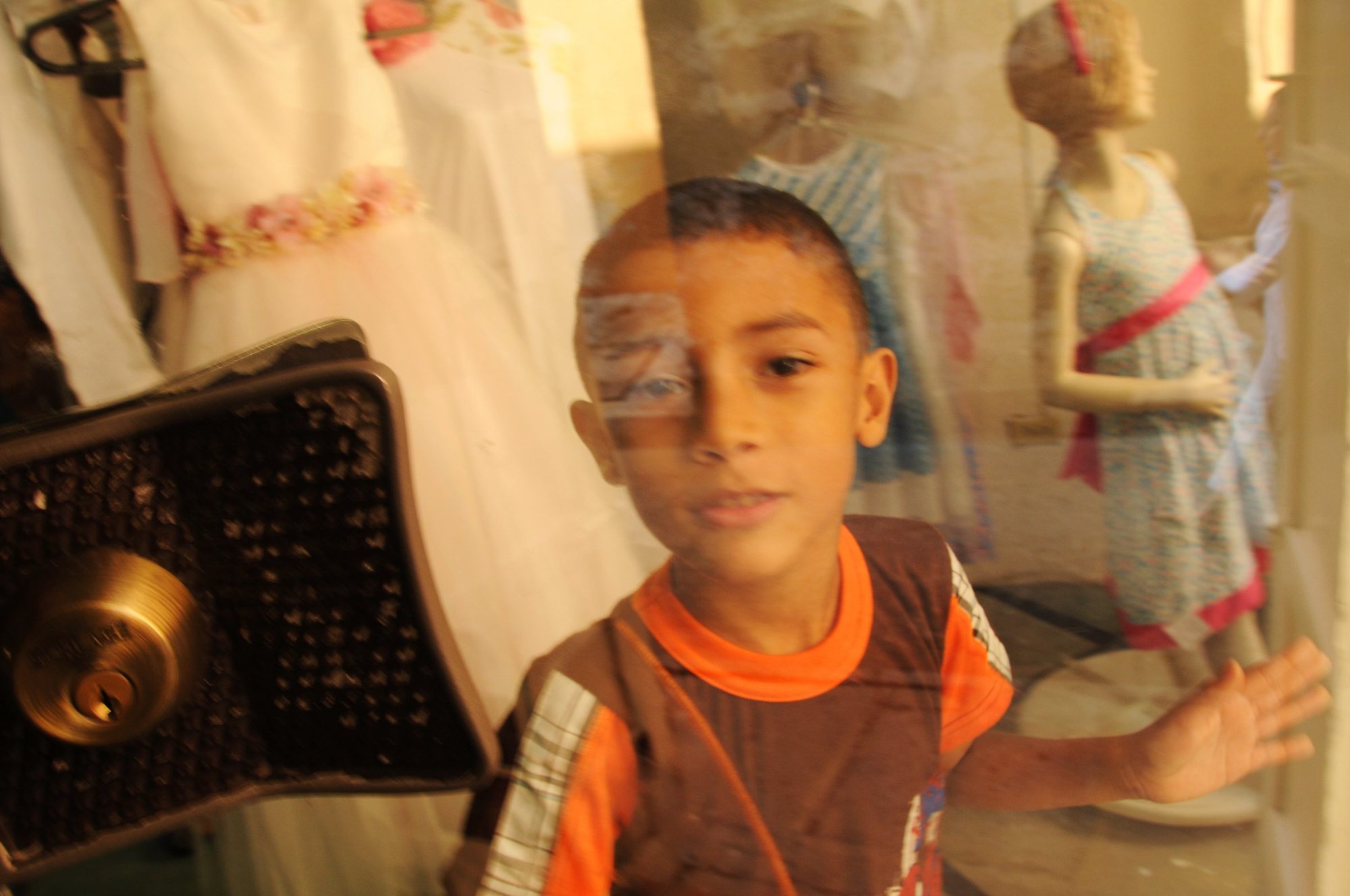 A boy seen through a shop window, dressed mannequins reflected across the glass, Cartagena, Colombia — photograph by Brian Walker