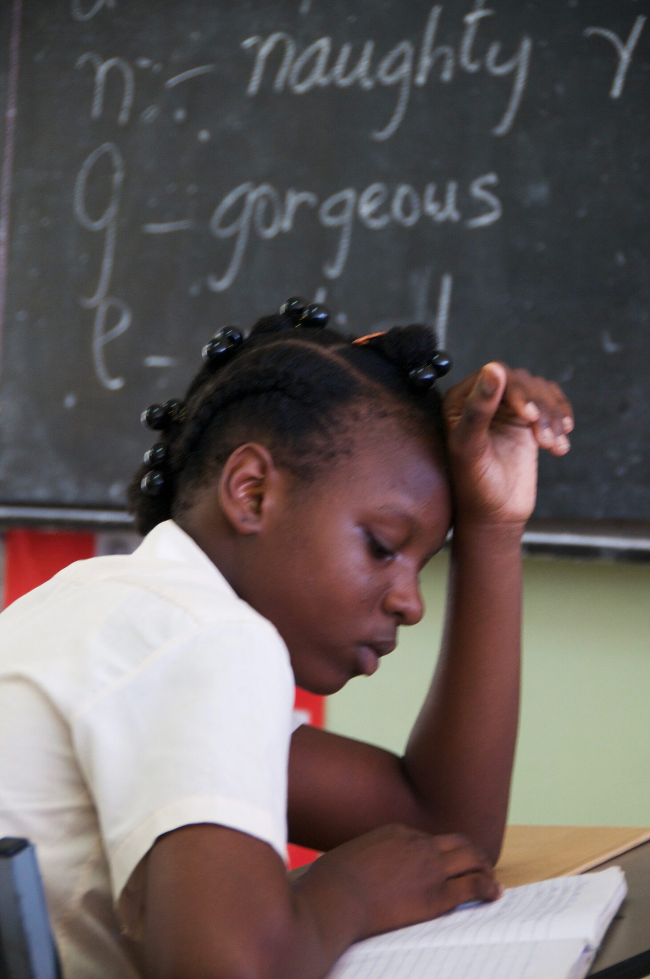 A schoolgirl reading at her desk in front of a blackboard covered in handwriting, Saint Vincent — photograph by Brian Walker