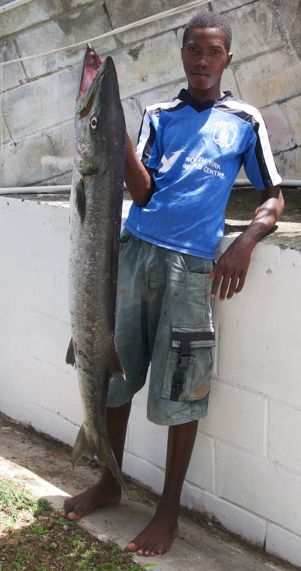 A young man in a blue football shirt leaning against a wall beside the large barracuda he has landed, Saint Vincent — photograph by Brian Walker