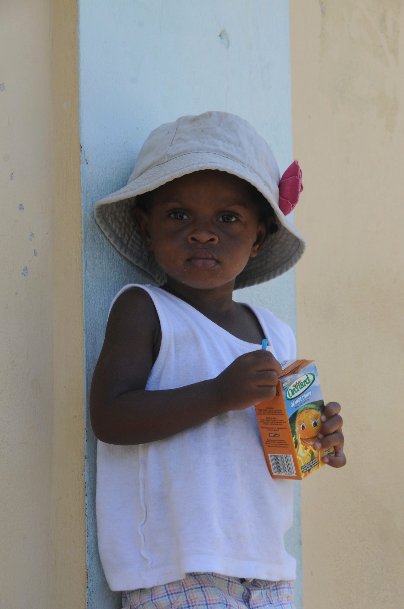 A small child in a white sun hat and vest holding a juice carton, standing in a painted doorway, Saint Vincent — photograph by Brian Walker