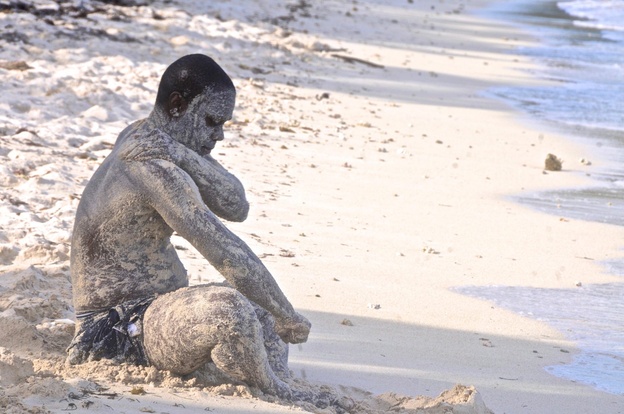 A boy crouched low on the beach, his body coated in wet sand, Saint Vincent — photograph by Brian Walker