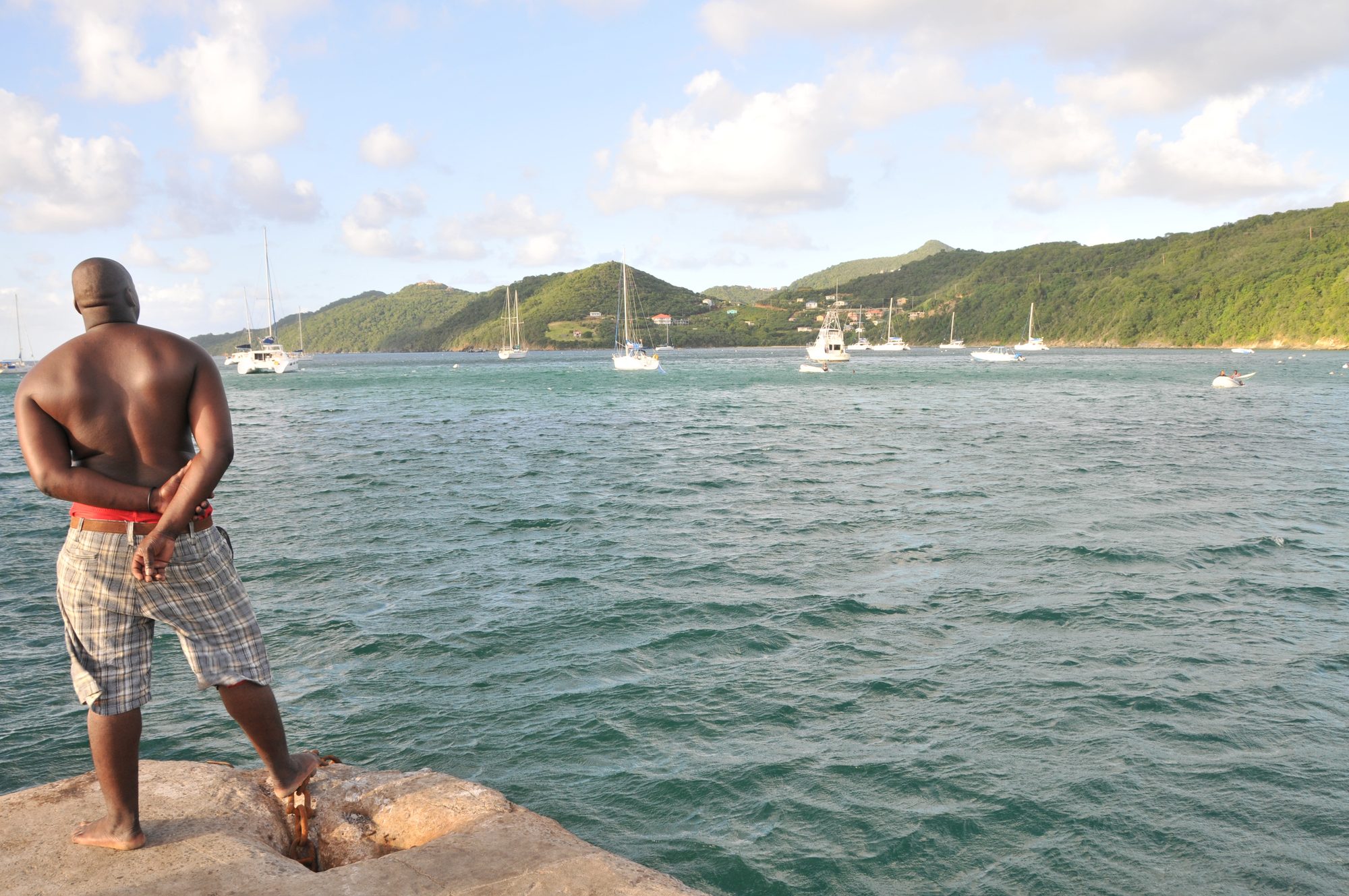 A man standing on rocks looking out across a harbour of moored sailboats, Saint Vincent — photograph by Brian Walker