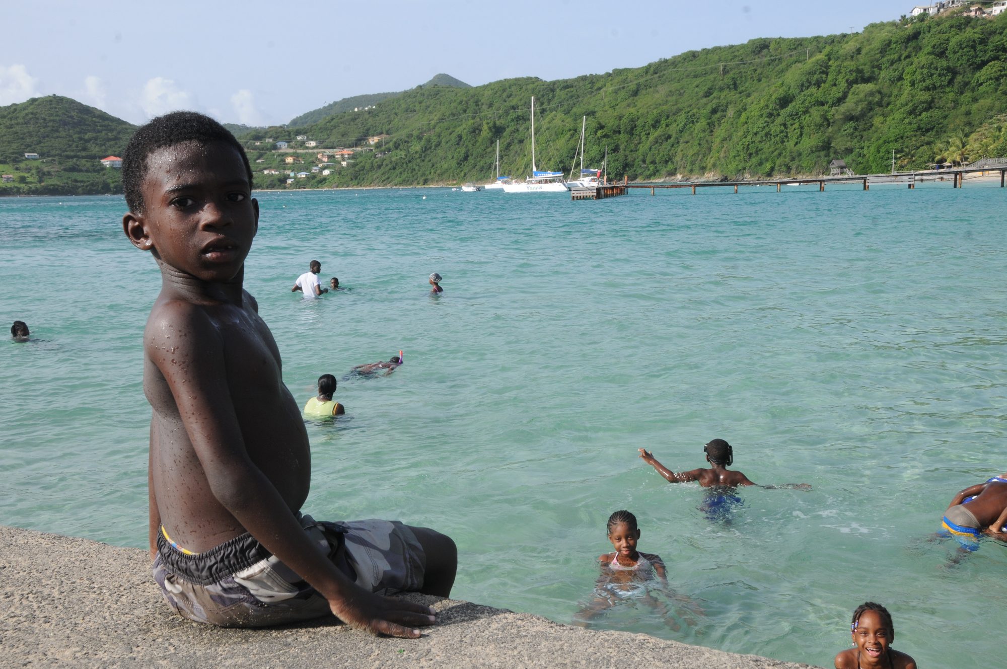 A boy sitting on a harbour wall watching other children swim in the bay, Saint Vincent — photograph by Brian Walker