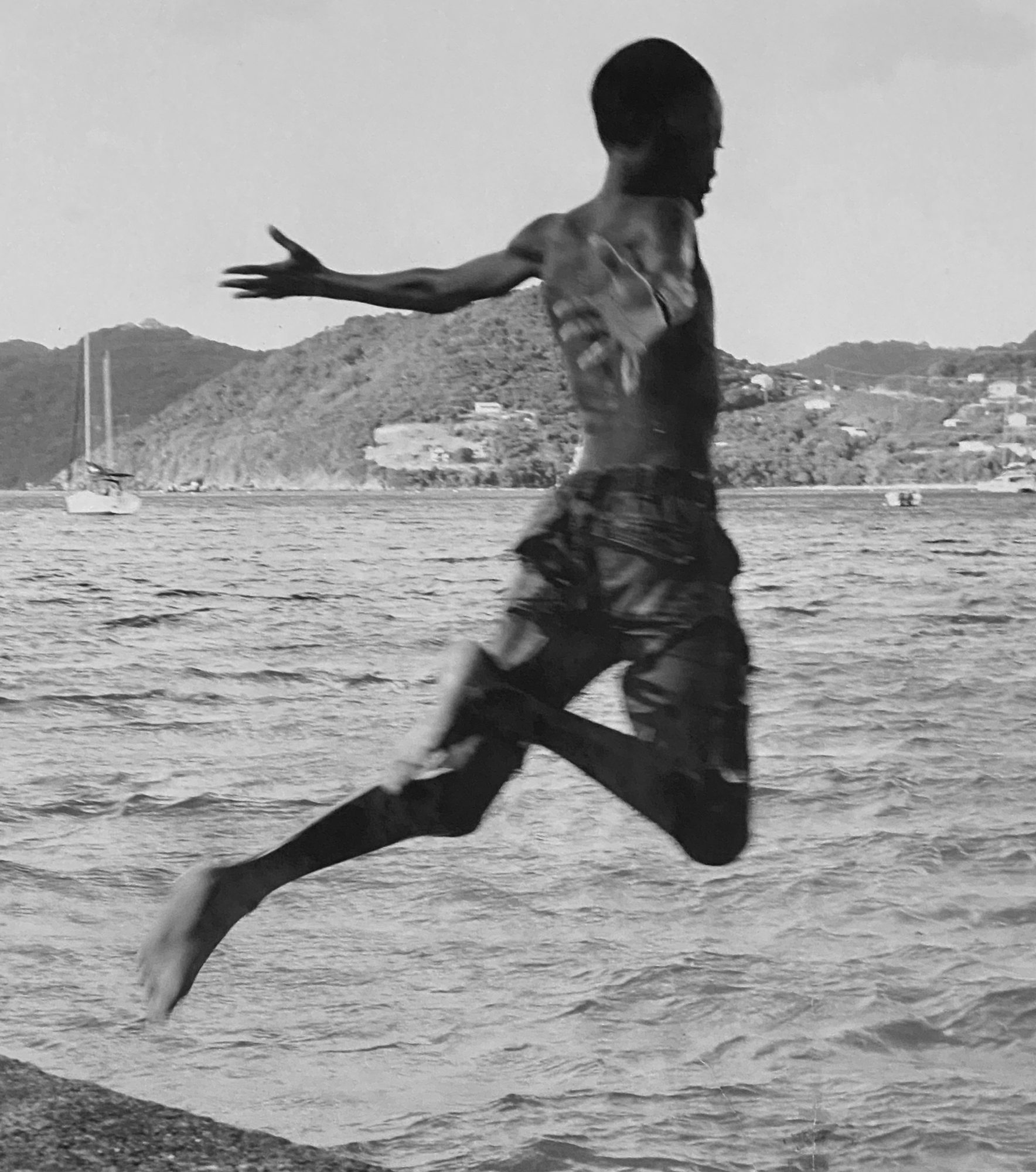 A boy caught mid-leap above the water, arms outstretched, a harbour behind him, black and white — photograph by Brian Walker