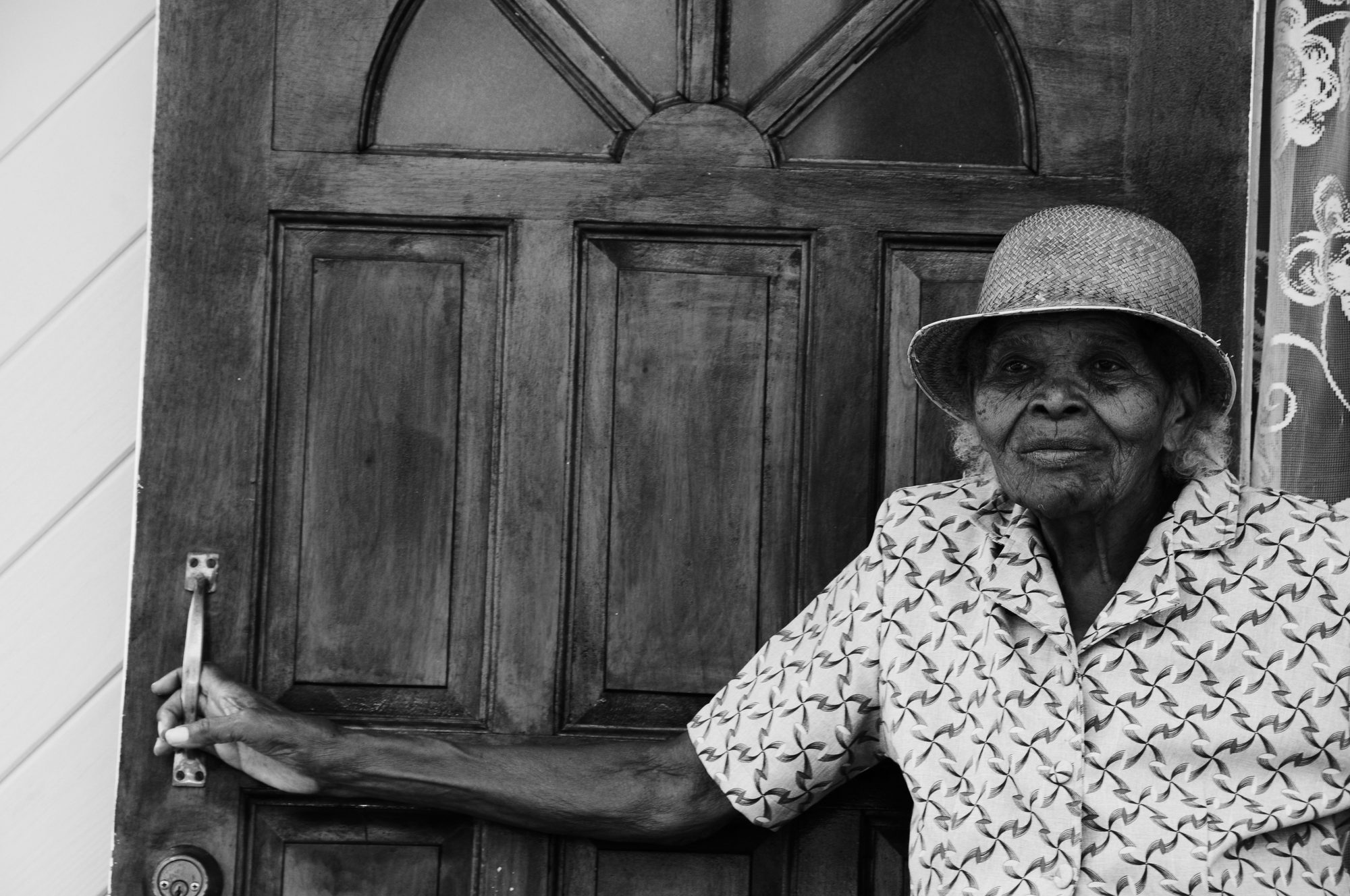 An elderly woman in a patterned dress and hat standing in an open wooden doorway, black and white — photograph by Brian Walker