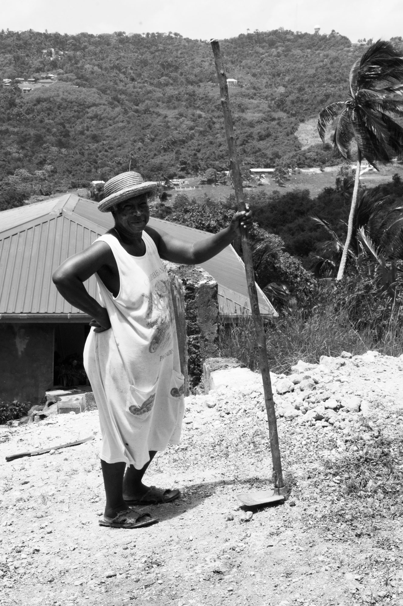 A woman in a sun hat standing on a hillside path, her hand resting on a long stick, black and white — photograph by Brian Walker