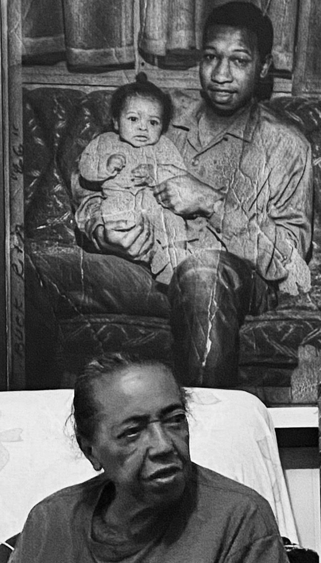 An older woman seated beneath a framed photograph of a man holding a baby, black and white — photograph by Brian Walker