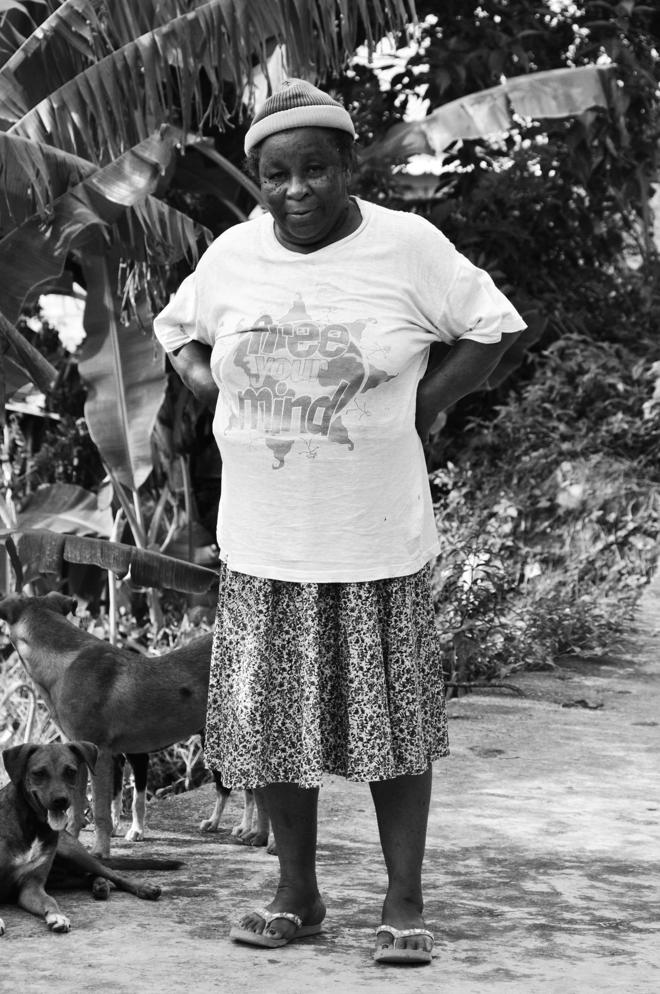 A woman standing outdoors among banana leaves, dogs resting at her feet, black and white — photograph by Brian Walker