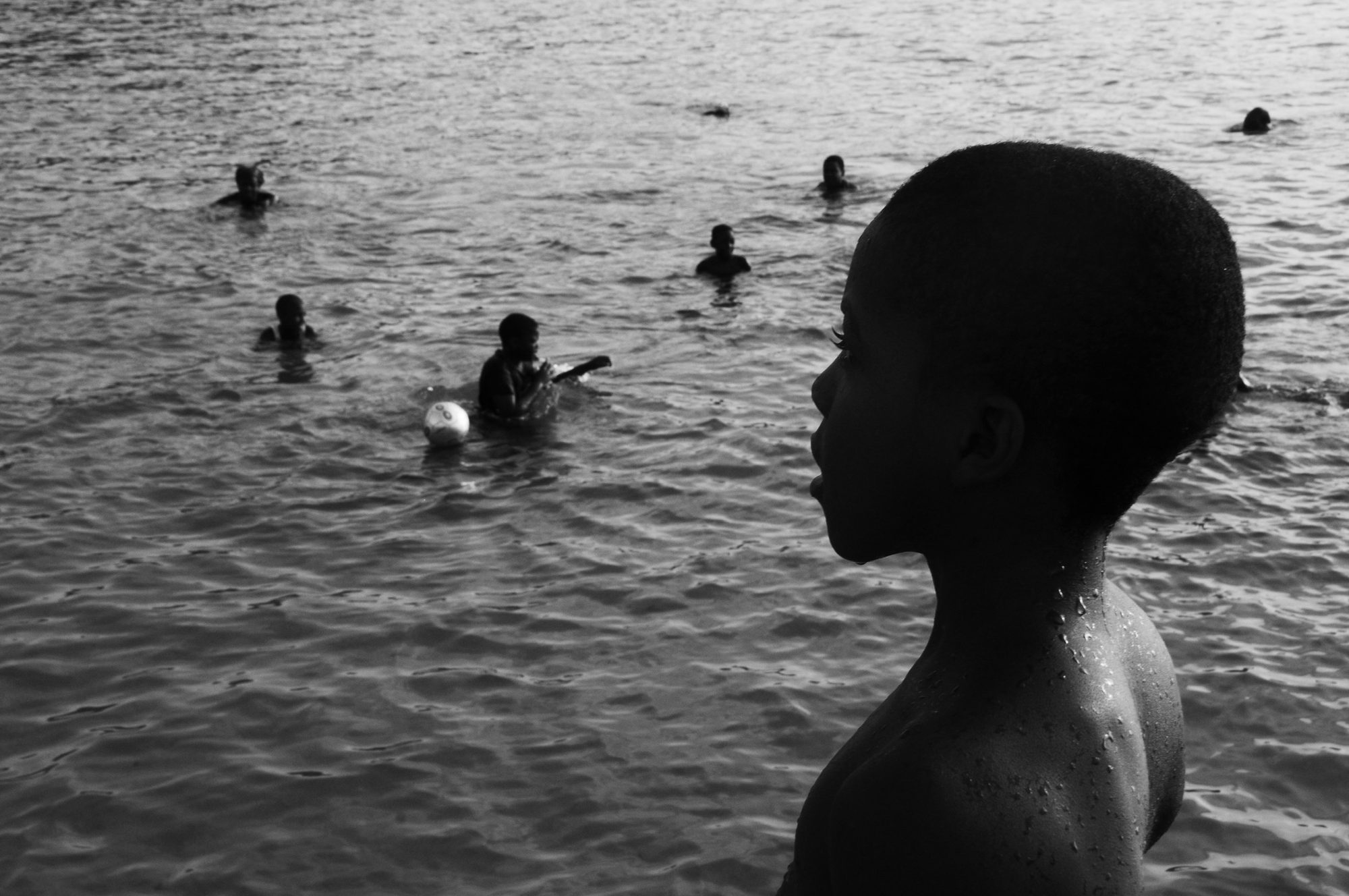 A boy in profile at the water's edge watching other children swimming, black and white — photograph by Brian Walker